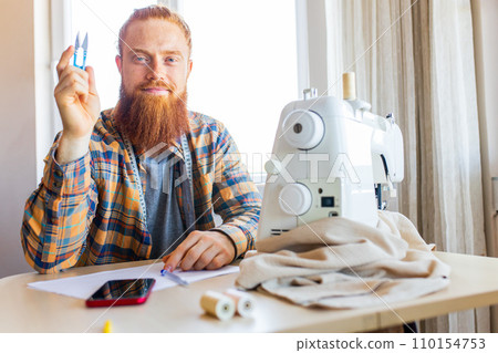 handsome redhaired man with long beard sews at a sewing machine at home studio handsome redhaired man with long beard sews at a sewing machine at home studio 110154753
