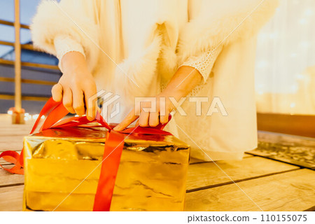 A woman in a white dress is holding a gold box with a red ribbon. She is wearing a crown on her head. The woman appears to be opening the gift box. A woman in a white dress is holding a gold box with a red ribbon. She is wearing a crown on her head. The woman appears to be opening the gift box. 110155075