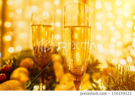 Two champagne glasses filled with champagne are placed on a table near to Christmas tree. The glasses are surrounded by lights, creating a festive atmosphere. Two champagne glasses filled with champagne are placed on a table near to Christmas tree. The glasses are surrounded by lights, creating a festive atmosphere. 110159121