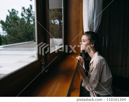 Portrait of young woman at home by window with wooden frame, autumn mood, cozy lifestyle, sleepy weather, relax on chair. 110161304