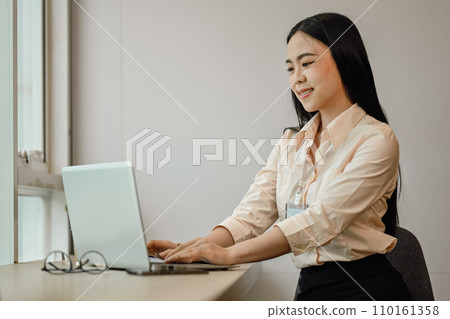 Attractive Asian female employee typing on laptop while working over presentation in bright office. Attractive Asian female employee typing on laptop while working over presentation in bright office. 110161358