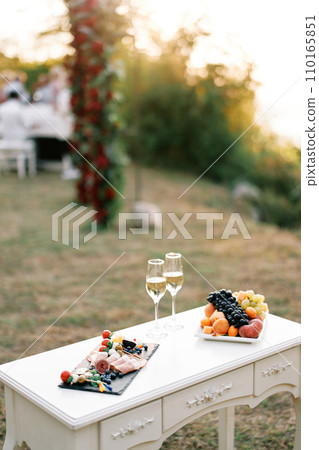 Table with two glasses of champagne and plates of festive treats stands in a clearing 110165851