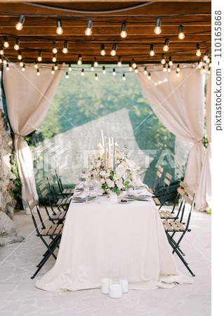 Laid table with bouquets of flowers stands on the terrace of an old house under a wooden canopy with curtains 110165868