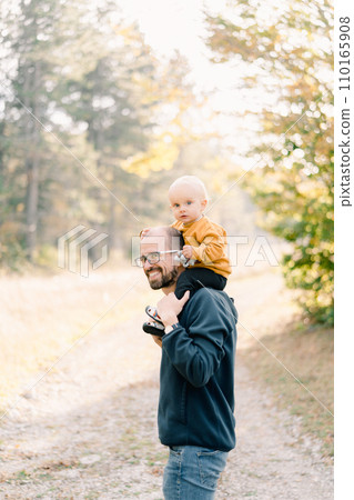 Smiling dad with a little boy on his shoulders stands half-turned in a clearing in the forest 110165908