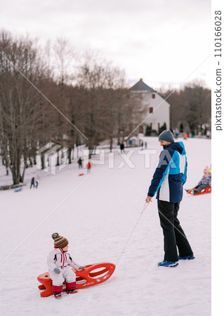 Dad is standing on a hill, holding a sled with a small child by the rope. Side view 110166028
