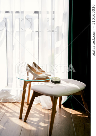 Bridal shoes stand near a box with wedding rings on a glass table near an easy chair in the room Bridal shoes stand near a box with wedding rings on a glass table near an easy chair in the room 110166036