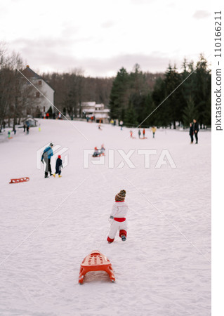 Small child in overalls drags a sled along a rope on a hill. Back view Small child in overalls drags a sled along a rope on a hill. Back view 110166211