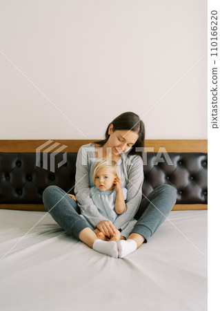 Mom hugs a little girl sitting behind her on the bed Mom hugs a little girl sitting behind her on the bed 110166220