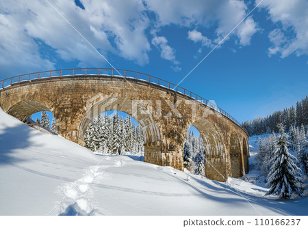 Stone viaduct (arch bridge) on railway through mountain snowy fir forest. Snow drifts  on wayside and hoarfrost on trees and electric line wires. 110166237