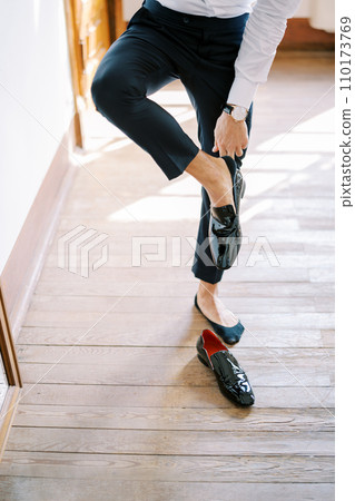 Groom puts on black patent leather shoes while leaning on the wall. Cropped. Faceless Groom puts on black patent leather shoes while leaning on the wall. Cropped. Faceless 110173769