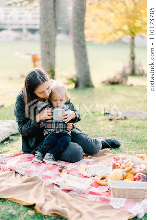 Mom with a little girl in her arms sits on a bedspread in an autumn park and gives her a drink from a mug 110173785