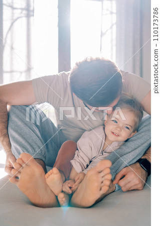 Dad kisses a little girl on the cheek while sitting with her on the bed with bare feet 110173786