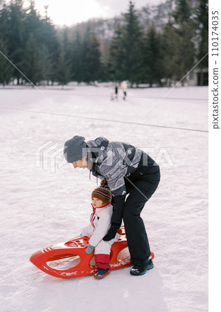 Mom puts a little girl on a sled on a snowy lawn 110174035