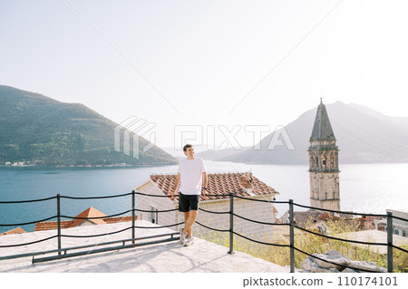 Smiling man leans on a rope fence on an observation deck near the church bell tower. Perast, Montenegro Smiling man leans on a rope fence on an observation deck near the church bell tower. Perast, Montenegro 110174101