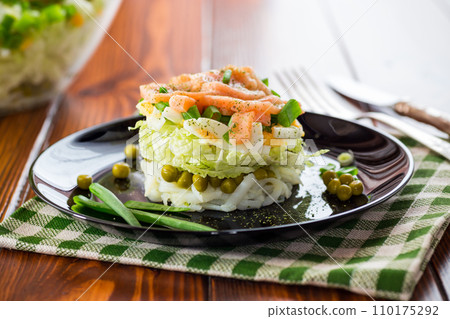 Layered salad of cabbage and other vegetables with pieces of red fish in a plate. 110175292