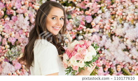 Pretty happy brunette woman with cute smile holding flower gift and posing on pink floral background Pretty happy brunette woman with cute smile holding flower gift and posing on pink floral background 110177610
