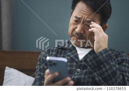 一名 50 多歲的穿著睡衣的男子一邊看著智慧型手機一邊做鬼臉 一名 50 多歲的穿著睡衣的男子一邊看著智慧型手機一邊做鬼臉 110178653