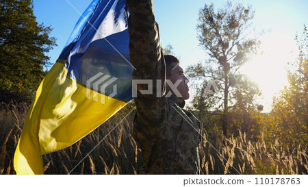 Military man in uniform stands with raised over head flag of Ukraine at countryside. Male ukrainian army soldier with lifted national banner in honor of victory against russian aggression. End of war. 110178763