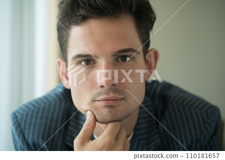 Close-up of young handsome guy in a white shirt, looks seriously and intently at the camera indoor 110181657
