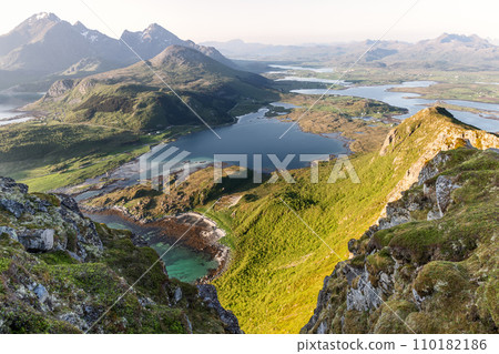 Verdant valleys and turquoise inlets of Lofoten bask in sunlight, with mountain backdrop 110182186