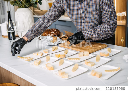 Catering Professional Arranging Cheese Plates for Service. Professional catering expert in black gloves places slices of cheese on individual plates for event, with backdrop of wine and fresh bread. 110183968