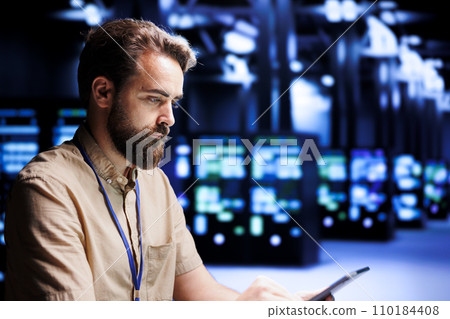 System administrator solving performance related issues in server cabinets. Expert utilizing tablet to identify operational problems causing high tech workplace infrastructure slowdown System administrator solving performance related issues in server cabinets. Expert utilizing tablet to identify operational problems causing high tech workplace infrastructure slowdown 110184408