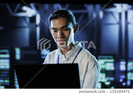 Asian man between server hub rows providing processing resources for businesses worldwide. System administrator fixing data center mainframes tasked with managing massive databases Asian man between server hub rows providing processing resources for businesses worldwide. System administrator fixing data center mainframes tasked with managing massive databases 110184471