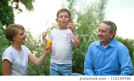 Father with mother looks at son blowing bubbles in fresh air during holidays Father with mother looks at son blowing bubbles in fresh air during holidays 110184596