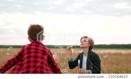Woman blows bubbles while energetic boy catches in field in cloudy weather Woman blows bubbles while energetic boy catches in field in cloudy weather 110184615