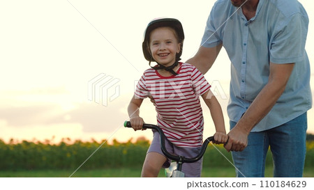 Smiling little girl learns to ride bicycle with positive father in summer evening Smiling little girl learns to ride bicycle with positive father in summer evening 110184629