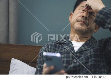 一名身穿睡衣的 50 多歲男子皺著眉頭看著智慧型手機 110184689