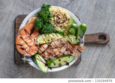 Breakfast is including Salmon steak, Chicken steak, Couscous, cut Avocado, Broccoli and cut Cucumber drizzled with Balsamic vinegar on Ceramic plate. Breakfast is including Salmon steak, Chicken steak, Couscous, cut Avocado, Broccoli and cut Cucumber drizzled with Balsamic vinegar on Ceramic plate. 110184976