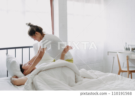 Funny couple wife try to wake lazy husband up in white bedroom. 110191568