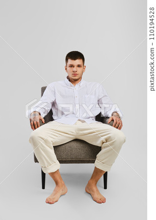Full length studio portrait of young barefoot man in white shirt and ivory trousers sitting in armchair 110194528