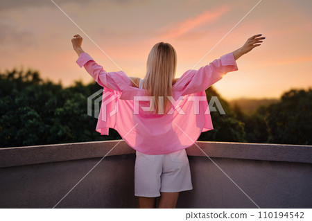 Back view of a woman standing with arms outstretched, embracing a colorful sunset 110194542
