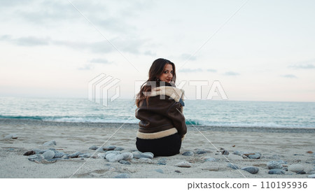 Girl seated on the beach near the ocean in overcast winter day Girl seated on the beach near the ocean in overcast winter day 110195936