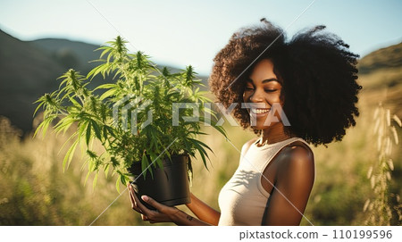 A woman holds a pot of flowering cannabis against a green field. A woman holds a pot of flowering cannabis against a green field. 110199596