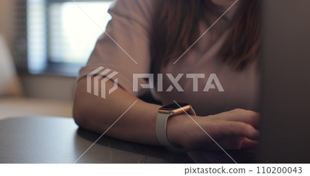 Woman fingers does double tap on smart watch for hands-free interaction. Young woman is typing on laptop at the work table at the office. Woman fingers does double tap on smart watch for hands-free interaction. Young woman is typing on laptop at the work table at the office. 110200043