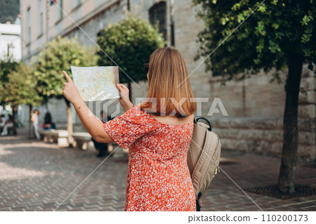 Happy Tourist girl with paper map walking in the city during vacation. Cheerful 30s woman in sunglasses traveling abroad in summer. Travel and active lifestyle concept, rear view 110200173