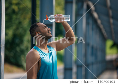 Young dark-skinned athlete pouring some refreshing water on himself after workout Young dark-skinned athlete pouring some refreshing water on himself after workout 110200303