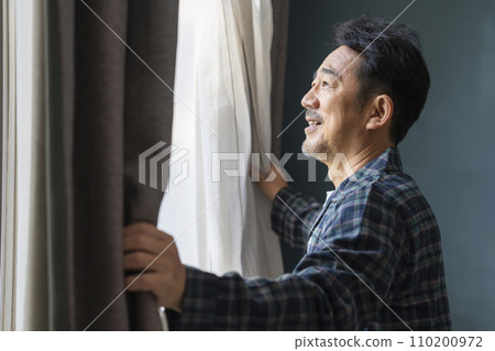 A man in his mid-50s wearing pajamas opening the curtains 110200972