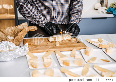Caterer Preparing Cheese Platters for an Event. Professional caterer arranges various types of cheese on plates, wearing gloves for hygiene while preparing for a gourmet event. Caterer Preparing Cheese Platters for an Event. Professional caterer arranges various types of cheese on plates, wearing gloves for hygiene while preparing for a gourmet event. 110203664