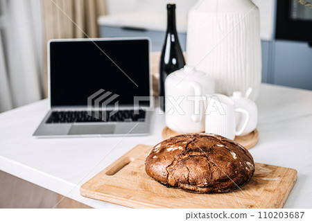 Homemade Bread on Kitchen Table with Laptop. Freshly baked homemade bread on a cutting board beside a laptop, depicting a cozy home baking scene possibly following an online recipe. 110203687