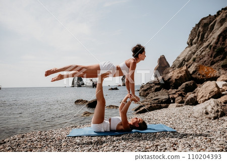 Woman sea yoga. Two Happy women meditating in yoga pose on the beach, ocean and rock mountains. Motivation and inspirational fit and exercising. Healthy lifestyle outdoors in nature, fitness concept. 110204393