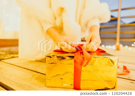 A woman in a white dress is holding a gold box with a red ribbon. She is wearing a crown on her head. The woman appears to be opening the gift box. 110204428