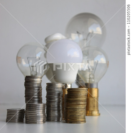 LED lamp and filament bulbs behind the piles of coins isolated on white. Concept photo for metaphoric increasing of energy saving LED lamp and filament bulbs behind the piles of coins isolated on white. Concept photo for metaphoric increasing of energy saving 110205506