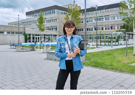 Middle-aged woman school teacher with digital tablet posing near school building 110205559