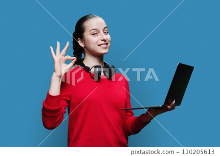 Teenage girl student in headphones using laptop showing hand gesture ok on blue background Teenage girl student in headphones using laptop showing hand gesture ok on blue background 110205613