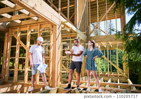 Man with detailed plan in hand showcases to couple the process of constructing wooden framed house. Investors inspecting their future dwelling. Concept of contemporary ecological construction. Man with detailed plan in hand showcases to couple the process of constructing wooden framed house. Investors inspecting their future dwelling. Concept of contemporary ecological construction. 110205717