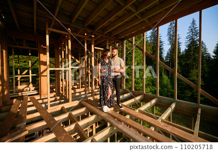 Man and woman inspecting their future wooden frame house nestled near the forest. Youthful couple at construction site in early morning. Concept of contemporary ecological construction. 110205718